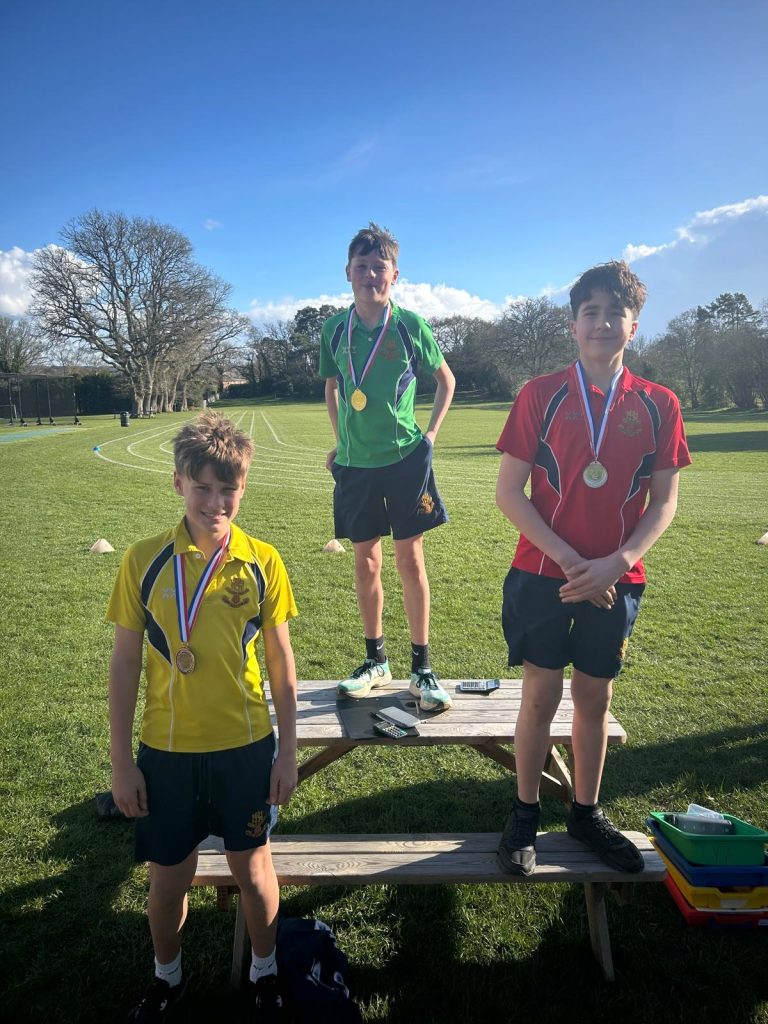 Three boys wearing medals stand on benches outdoors in a sports field; each wears a different coloured shirt—yellow, green, and red—under a sunny sky.
