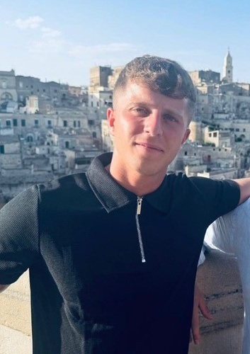 A young man in a black zip-up top stands outdoors in front of an old cityscape with stone buildings under a clear sky, as crowds gather nearby for the celebrity football match.