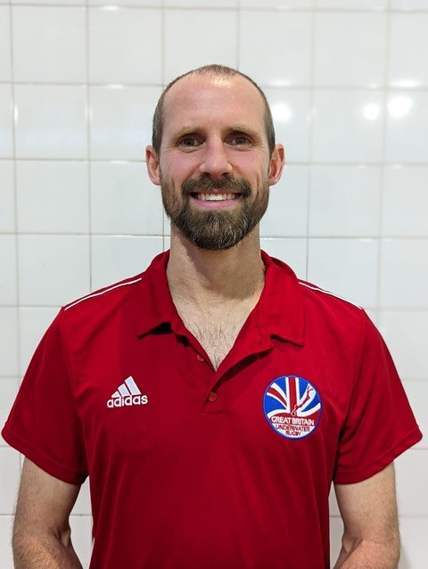A man with a beard stands in front of a tiled wall, wearing a red Adidas polo shirt with a Great Britain team logo.