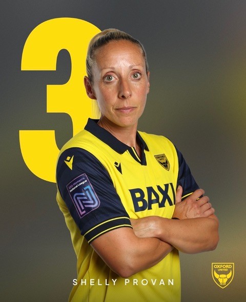 A woman in a yellow Oxford United football shirt stands with arms folded, looking at the camera.