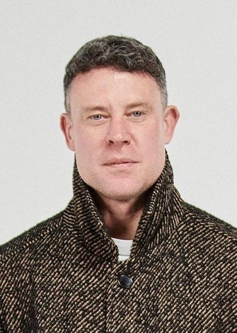 A man with short, dark hair wearing a patterned wool coat with the collar turned up, posing as if ready for a celebrity football match against a plain light background.