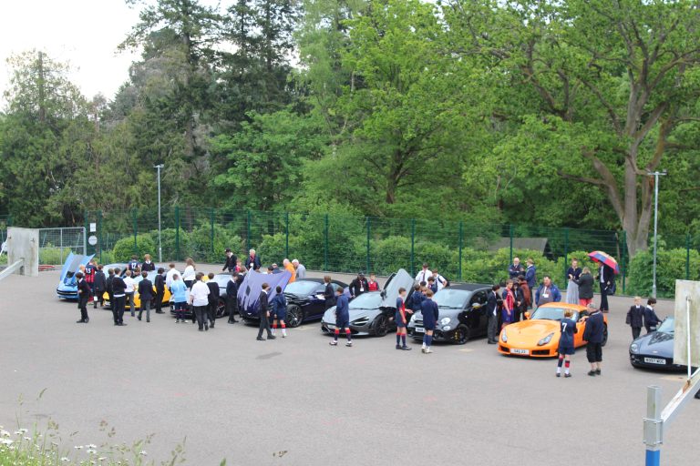 A group of students and adults gather around several sports cars.