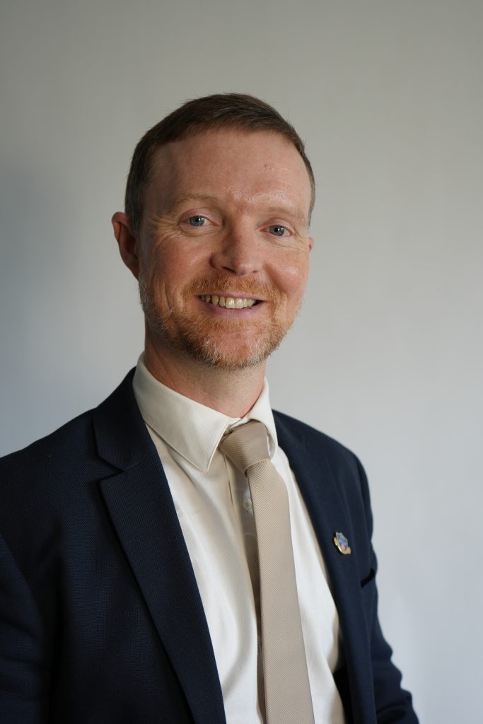 A man in a suit and beige tie stands against a plain light background, smiling at the camera.
