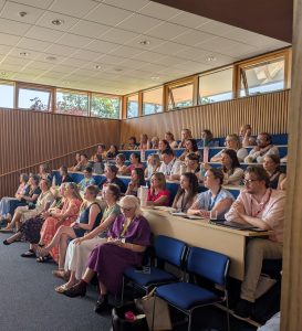 A group of adults sit in tiered rows of a lecture theatre, attentively listening to a presentation.