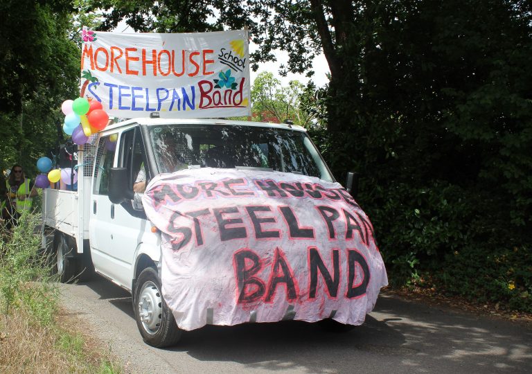 A white lorry decorated with colourful banners and balloons displays the text “More House Steel Pan Band” as it drives down a tree-lined road.