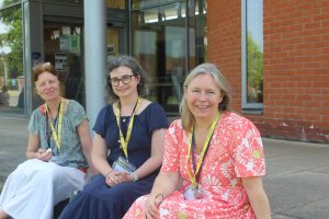 Three women wearing lanyards sit outside a brick building, smiling at the camera. One holds a notepad, and sunlight brightens the scene.