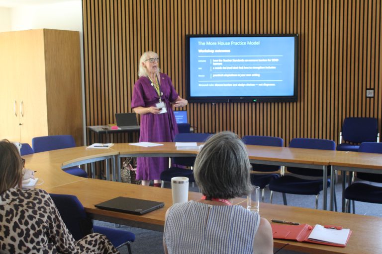 A woman in a purple dress presents in a conference room to a small group. A screen behind her displays The More House Practice Model workshop outcomes.