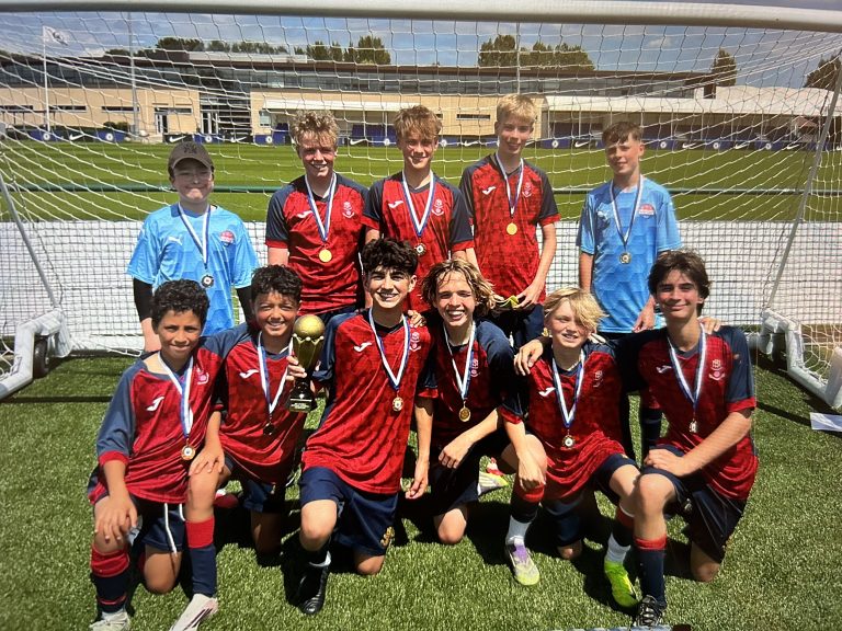 Eleven boys in football kits pose in front of a goal on a pitch, wearing medals, with one holding a trophy.