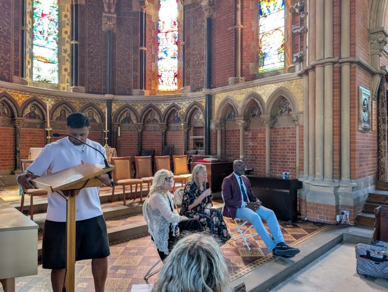 A person reads at a lectern in a church whilst three people sit and listen on chairs; stained glass windows and ornate walls are visible in the background.