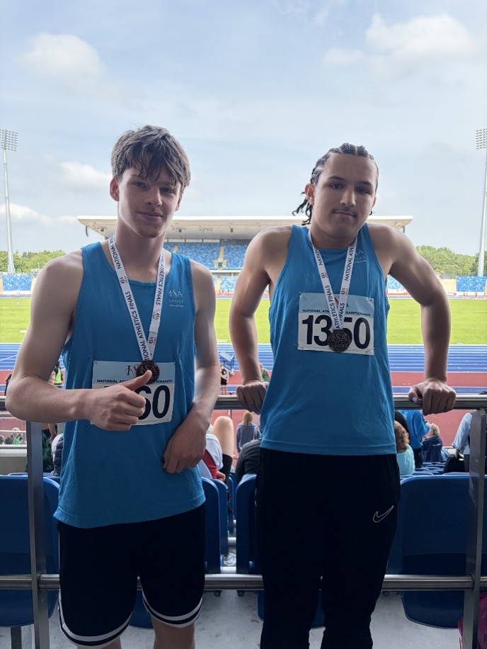 Two young athletes wearing blue jerseys and medals stand side by side in a stadium with a track and field in the background.